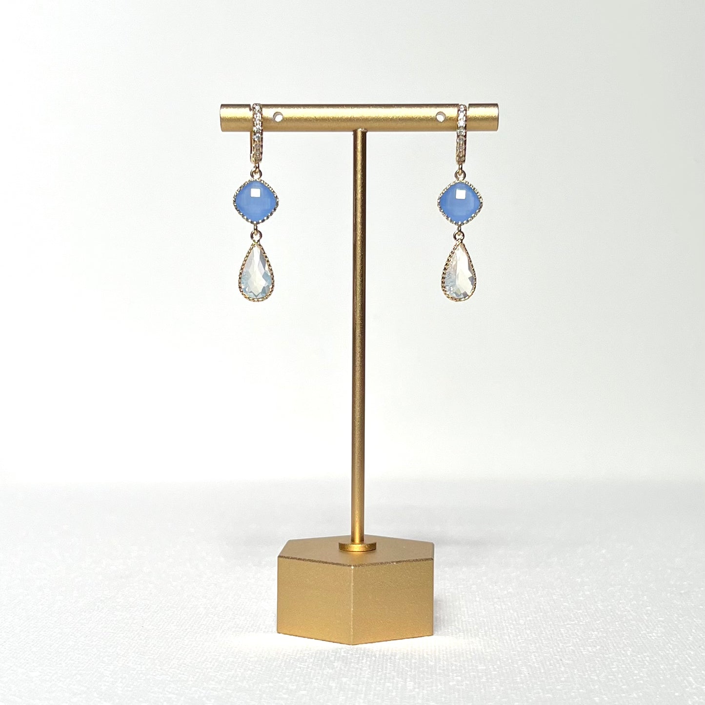 This earring set features a blue glass fancy prong set bezel charm with a milky white glass teardrop shaped prong set bezel hung by a gold filled CZ crested huggie style hoop.&nbsp;