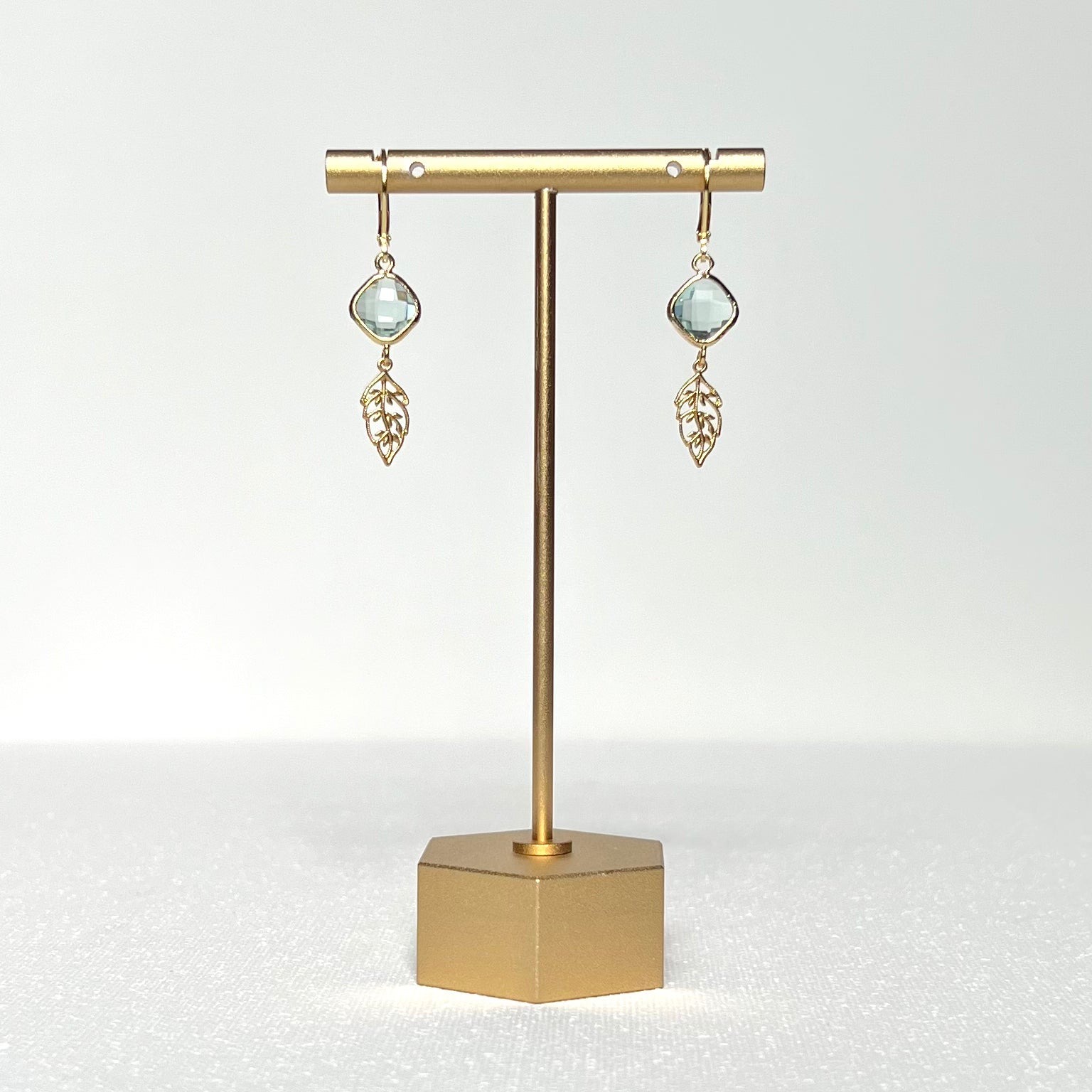 This earring set features a pale green glass charm in a gold bezel with a gold leaf, hung on a French lever back style hook.&nbsp;